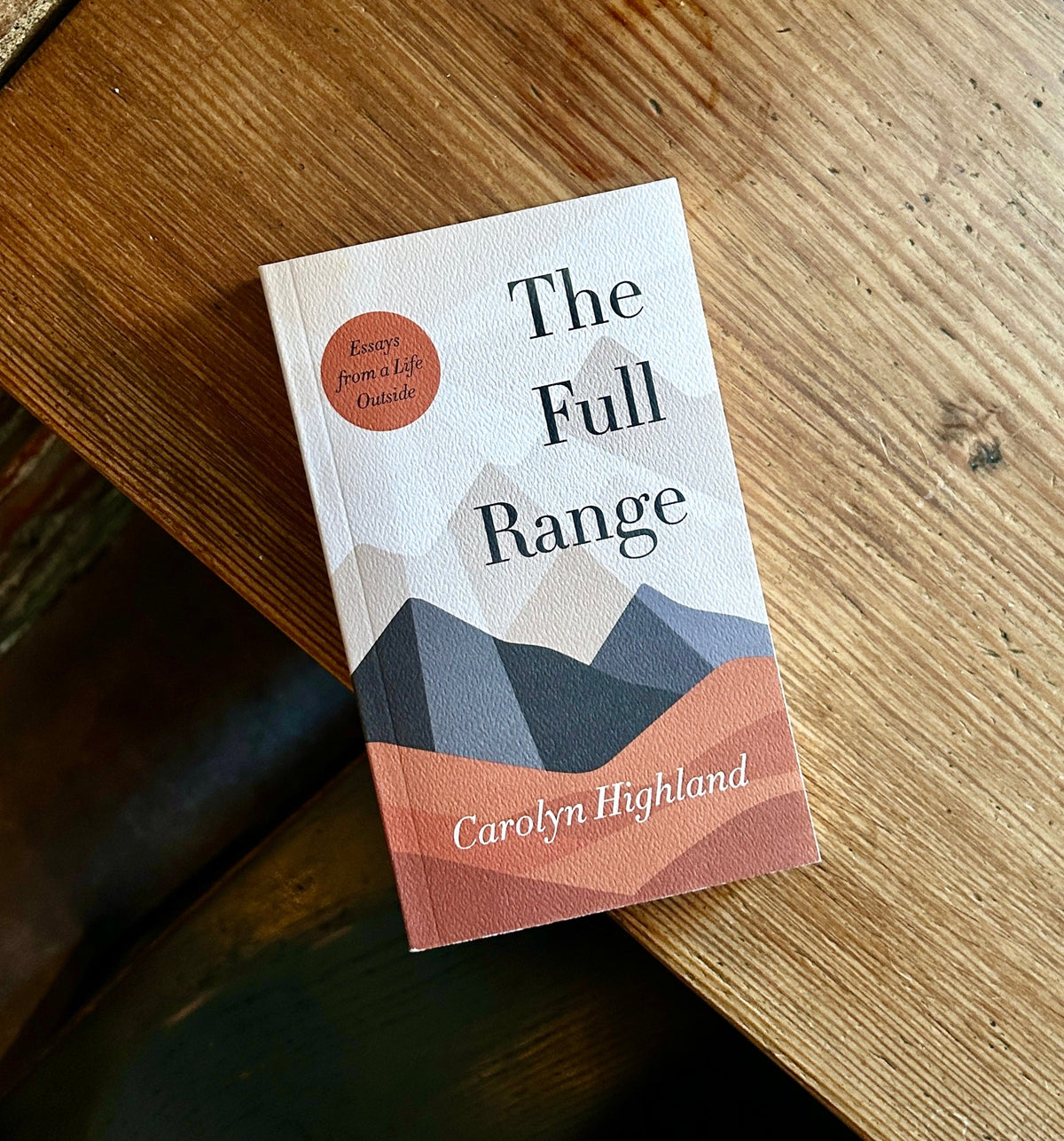 A photograph of The Full Range: Essays from a Life Outside by Carolyn Highland. The book is pictured at an angle on the edge of a rustic wooden table.